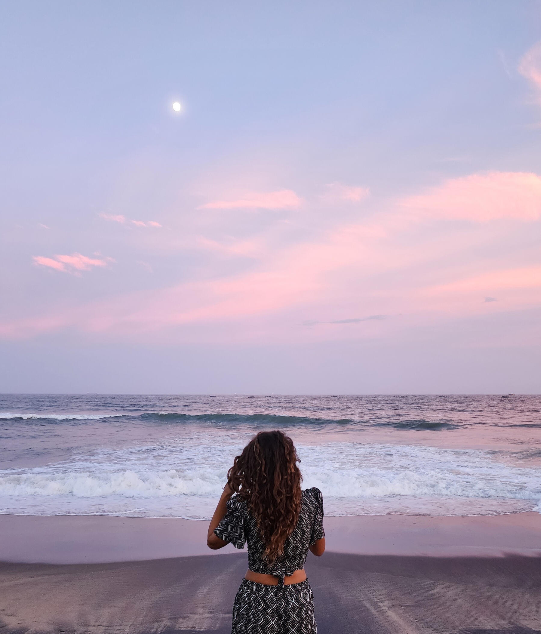 A Grounded Pause by the Ocean Samara standing by the ocean at sunset, facing the waves under a soft pink and purple sky.